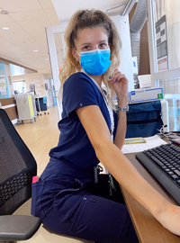 Selma Brkic wearing navy scrubs and blue surgical mask sits at desk in hospital hallway